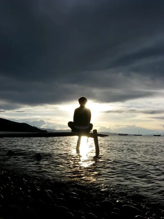 A silhouetted figure sitting cross-legged at the end of a jetty, meditating as the sun sets over water