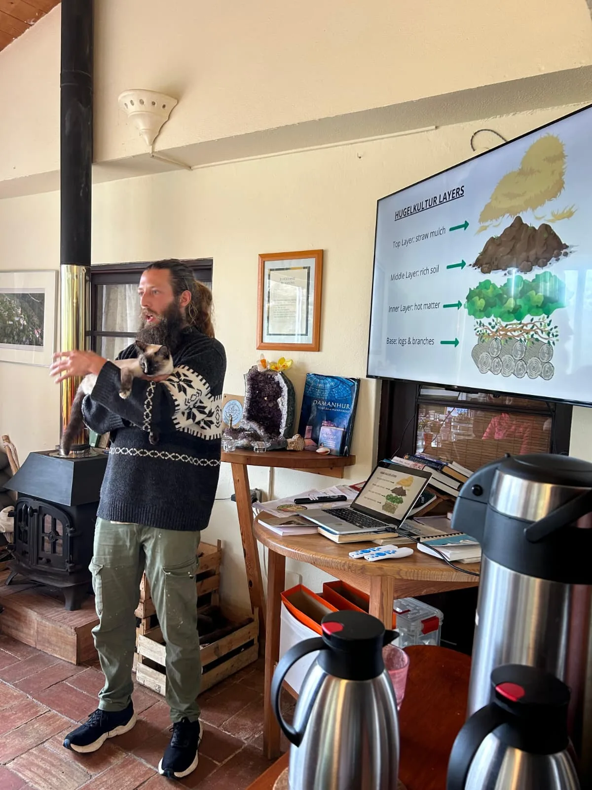 Benjy teaching a hugelkultur workshop beside a diagram screen, a cat resting in his arms